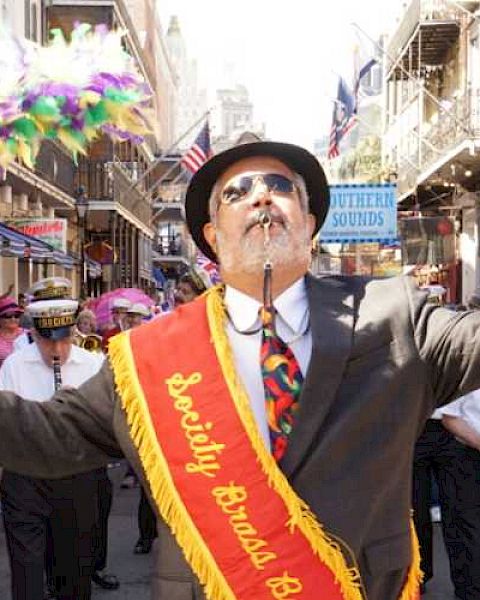 A man leads a brass band parade through a lively street crowd.